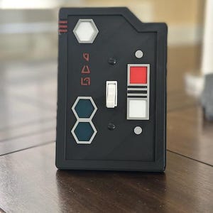 May include: A black light switch cover with a white toggle switch and a red button. The cover has a hexagonal design with the word "504" printed on it.