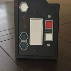 May include: A black plastic light switch cover with a white toggle switch, a red button, and a hexagonal button with a green design. The cover has the text "545" printed on it.