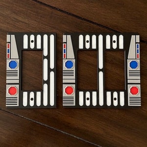 May include: Two black and white light switch covers with a geometric design. Each cover has a rectangular opening, with a red and blue circle and a silver accent. The covers are on a dark wood surface.