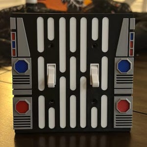 May include: A black light switch cover with a white Star Wars design. The cover has two toggle switches and red and blue accents.