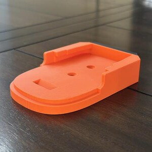 May include: Orange plastic battery charging stand with a black trim. The stand has two small holes and a slot for the battery.