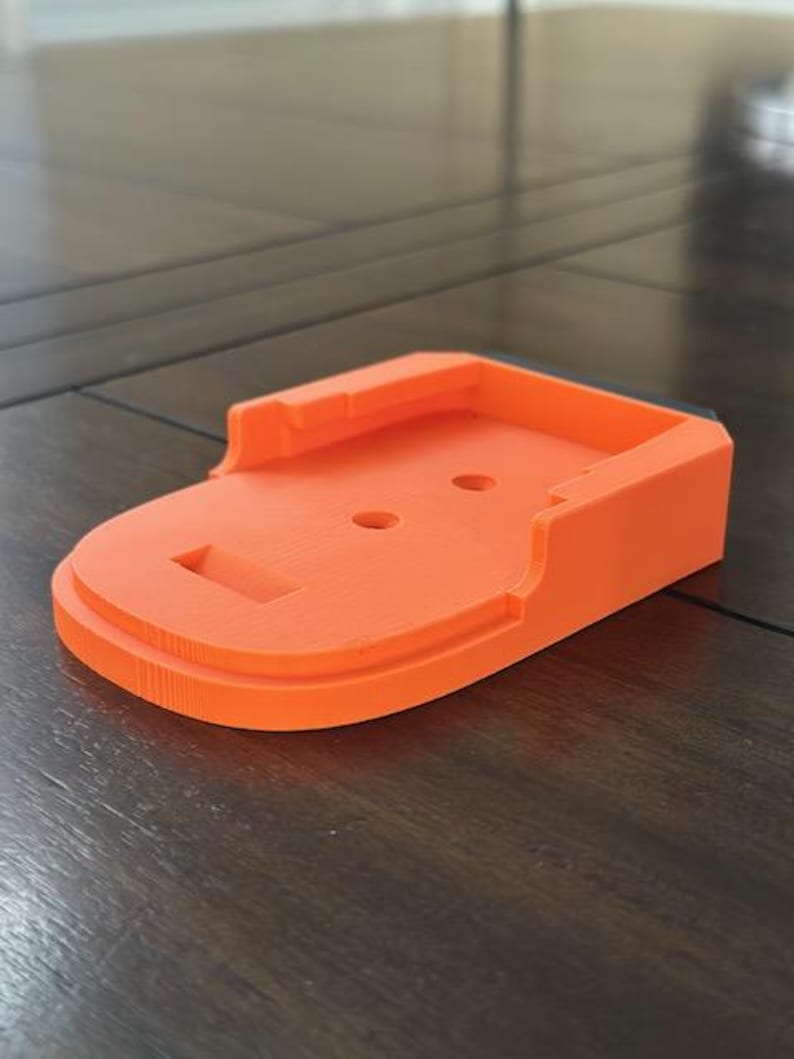 May include: Orange plastic battery charging stand with a black trim. The stand has two small holes and a slot for the battery.