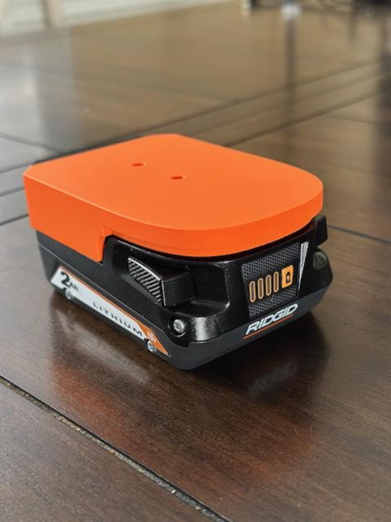 May include: An orange and black Ridgid 18V lithium-ion battery with a 2Ah capacity. The battery has a digital display that shows the charge level.