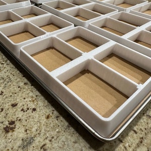 May include: A white baking pan with multiple square compartments. The pan is made of metal and has a brown, cardboard-like liner.