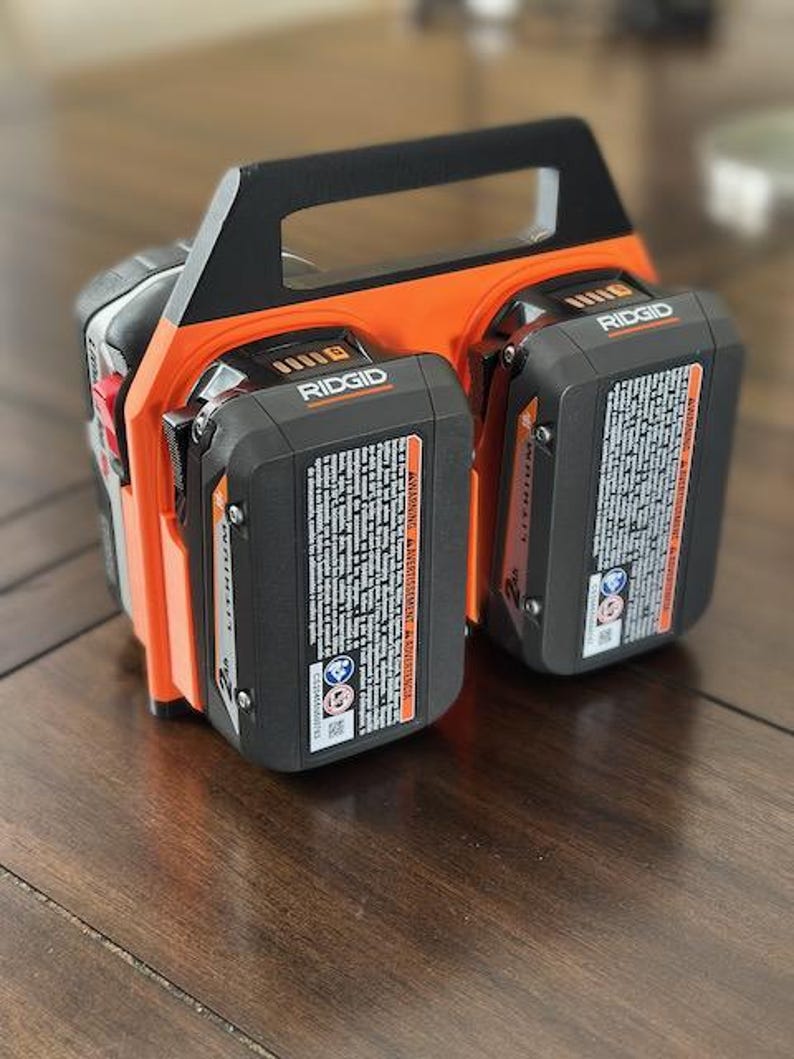 Pode incluir: Um carregador de bateria preto e laranja com duas baterias RIDGID pretas. O carregador tem uma al&ccedil;a e um cabo de alimenta&ccedil;&atilde;o.