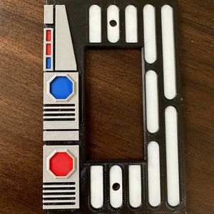 May include: A black, white, gray, and red rectangular decorative frame with geometric shapes. The frame has a central opening and features a series of vertical white bars on a black background. The frame also has a series of rectangular and octagonal shapes with red and blue accents.