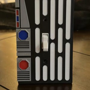 May include: A black and white light switch cover with a Star Wars design. The cover has a white rectangular panel with a series of vertical lines and a toggle switch. The cover also has a gray panel with a red and blue button.