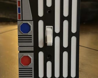 May include: A black and white light switch cover with a Star Wars design. The cover has a white rectangular panel with a series of vertical lines and a toggle switch. The cover also has a gray panel with a red and blue button.