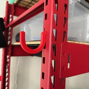 May include: Red metal shelving unit with a red plastic hook attached to the side. The hook is shaped like a 'J' and is designed to hold items.