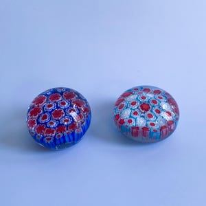 May include: Two glass paperweights with floral patterns. One is mainly blue with red flowers, the other is white with red and blue flowers. The paperweights are round and sit on a light blue surface.