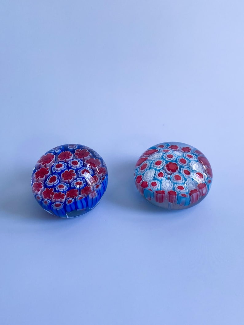 May include: Two glass paperweights with floral patterns. One is mainly blue with red flowers, the other is white with red and blue flowers. The paperweights are round and sit on a light blue surface.