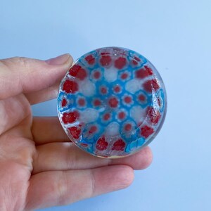 May include: A circular glass object with a red, white, and blue pattern. The design features red and white shapes surrounded by blue. The object is held in a hand against a light blue backdrop.