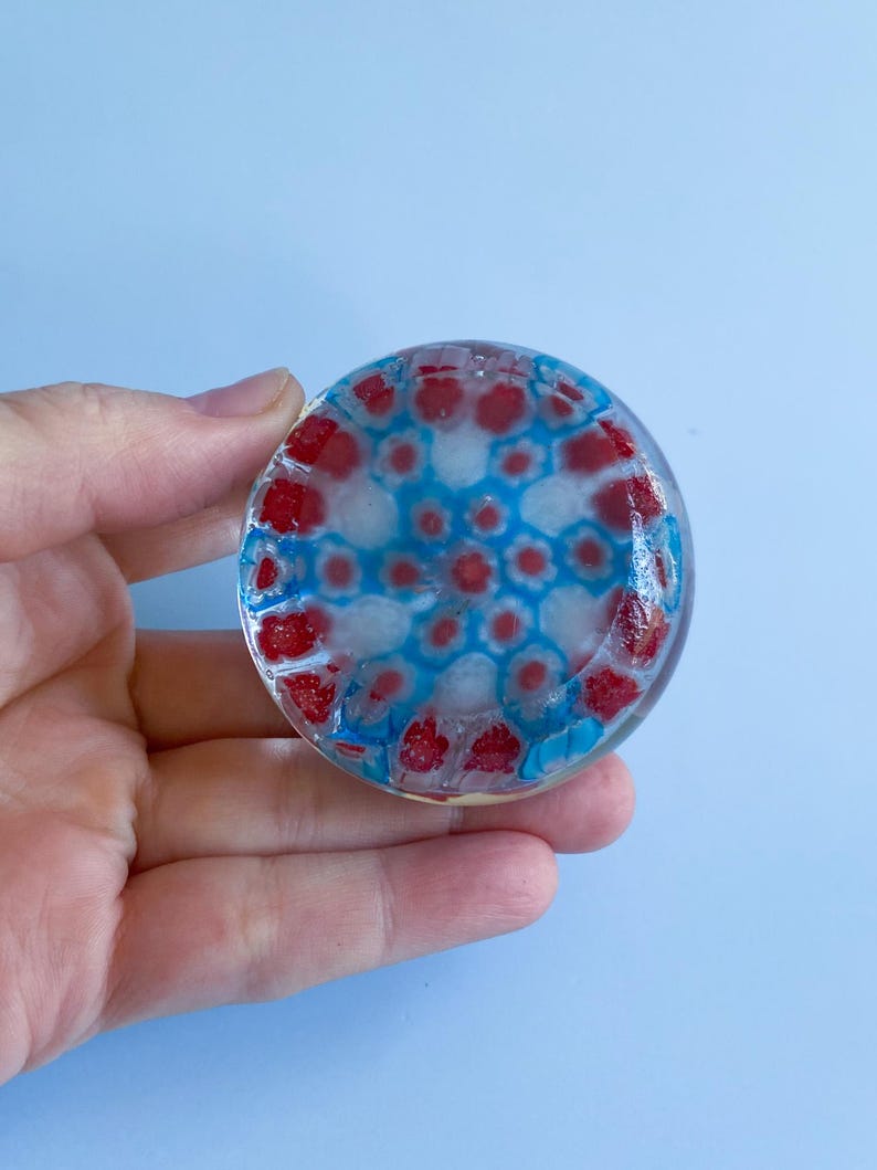 May include: A circular glass object with a red, white, and blue pattern. The design features red and white shapes surrounded by blue. The object is held in a hand against a light blue backdrop.