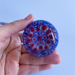 May include: A round, blue glass paperweight with a floral pattern in red and white. The paperweight is held in a hand against a light blue background. The glass has a glossy finish and the floral design is visible throughout.
