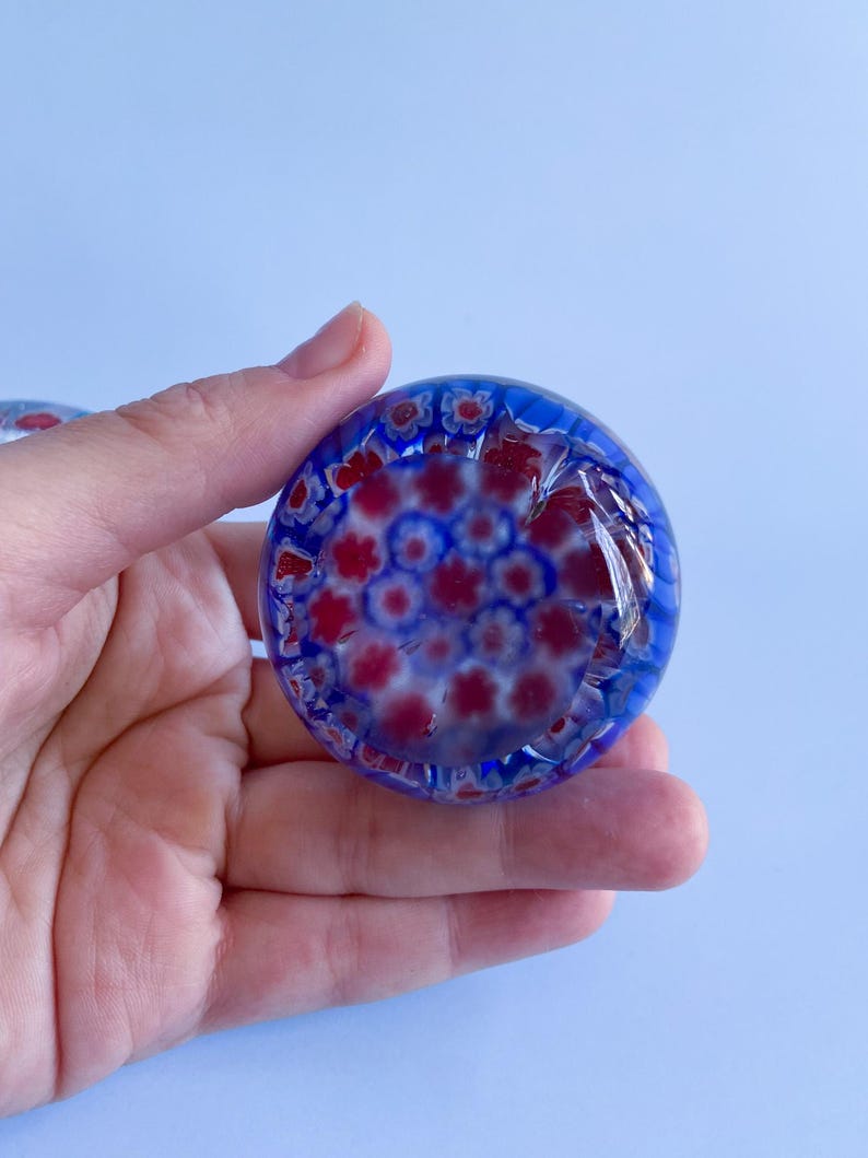 May include: A round, blue glass paperweight with a floral pattern in red and white. The paperweight is held in a hand against a light blue background. The glass has a glossy finish and the floral design is visible throughout.