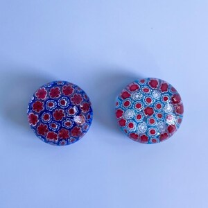 May include: Two round glass paperweights with floral patterns. One has a blue background with red flowers, the other has a light blue background with red and white flowers. The paperweights are decorative and colourful.