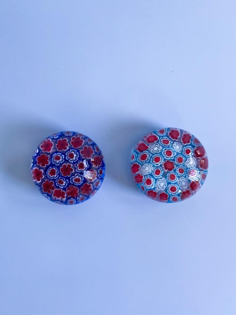 May include: Two round glass paperweights with floral patterns. One has a blue background with red flowers, the other has a light blue background with red and white flowers. The paperweights are decorative and colourful.