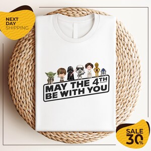 A white t-shirt with a graphic design featuring characters from the Star Wars franchise, along with the text "May the 4th be with you".