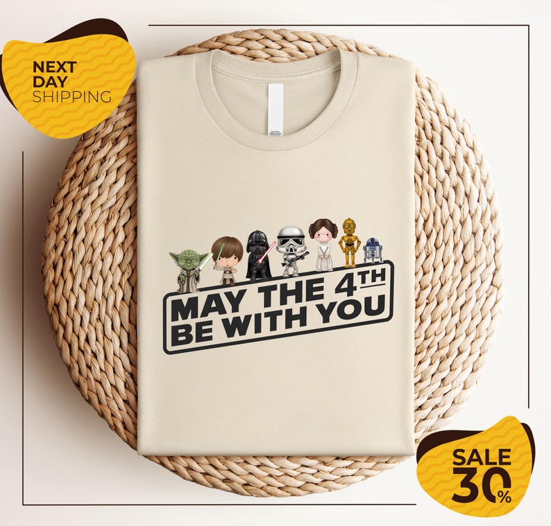 A beige t-shirt with a graphic design featuring characters from the Star Wars franchise, along with the text "May the 4th be with you".