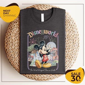 A t-shirt featuring a Mickey Mouse graphic with various Disney-themed elements, such as a castle, fireworks, and a sign that says "Disneyworld". The t-shirt is displayed on a woven placemat, and there are two yellow price tags in the image.