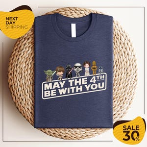 A navy blue t-shirt with a graphic design featuring characters from the Star Wars franchise, along with the text "May the 4th be with you". The t-shirt is displayed on a woven basket, and there are two yellow sale signs indicating a 30% discount.
