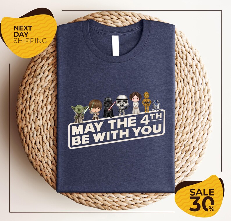 A navy blue t-shirt with a graphic design featuring characters from the Star Wars franchise, along with the text "May the 4th be with you". The t-shirt is displayed on a woven basket, and there are two yellow sale signs indicating a 30% discount.