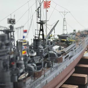 Puede incluir: Maqueta detallada de un buque de guerra japonés, con casco gris y parte inferior roja. El modelo incluye mástiles, aparejos y una bandera japonesa. El barco está montado sobre soportes de madera.