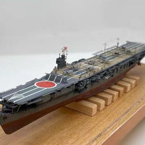 May include: A detailed model of a Japanese aircraft carrier, featuring a red circle emblem on the deck. The model is painted in shades of gray, brown, and red, and is mounted on a wooden base. Several miniature airplanes are visible on the deck.