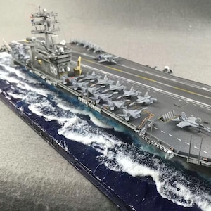 Könnte beinhalten: Detailliertes Modell eines Flugzeugträgers mit zahlreichen Kampfflugzeugen auf dem Deck. Das Modell hat ein graues Flugdeck, einen Aufbau mit der Nummer 66 und eine blau-weiße Ozeanbasis. Das Modell ist eine Miniaturreplik.