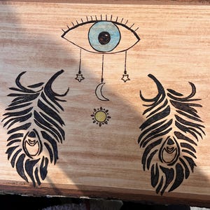 May include: A wooden board with a blue eye, two black feathers, a crescent moon, a sun, and two stars. The eye is surrounded by eyelashes and has a white pupil. The feathers are detailed with black lines and have a white eye in the center. The moon is a crescent shape and the sun is a circle with rays. The stars are five-pointed.