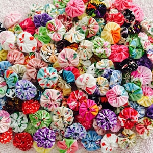 May include: A collection of colorful fabric flower embellishments. The flowers are made of various fabrics and patterns, including floral prints, polka dots, and stripes. The flowers are all different sizes and shapes, and they are arranged in a random pattern.