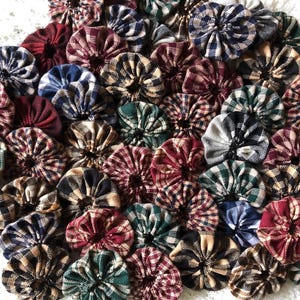 May include: A collection of numerous fabric yo-yos, which are small, circular, gathered textile ornaments. These pieces feature a variety of plaid and checkered patterns in colors including red, green, blue, black, white, and tan, arranged together.