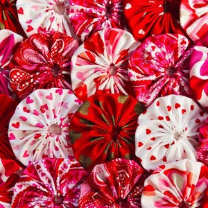 May include: A close-up of a collection of red, pink, and white fabric rosettes with a heart pattern. The rosettes are arranged in a random pattern and are all different sizes.