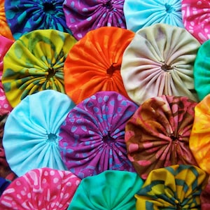 May include: A close-up of a collection of colorful fabric circles. The circles are in various shades of pink, orange, yellow, green, blue, and purple. Each circle is gathered in the center, creating a textured, flower-like appearance.
