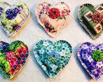 Puede incluir: Seis adornos de corazón hechos a mano de tela con diferentes patrones florales y colores. Cada corazón tiene un botón en el centro y está bordeado con costuras blancas.