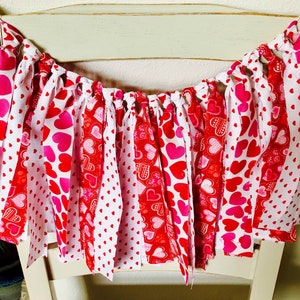 May include: A red and white fabric banner with hearts printed on it. The banner is strung on a white chair.