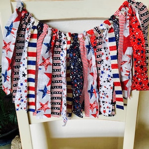 May include: A red, white, and blue fabric banner with stars and stripes. The banner is made of strips of fabric sewn together and is hanging from a white chair.
