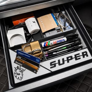 Op de afbeelding: Een auto-lade-organizer gevuld met diverse items. Inclusief pennen, sleutels, een bril, een slot, kaarten en een container met een bliksemgrafiek. Het woord "SUPER" wordt weergegeven op een witte achtergrond.