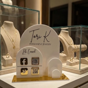 May include: A display for Tori K Permanent Jewelry featuring a white and gold design. The display includes the brand's logo, social media icons, and QR codes. Jewelry pieces are showcased in glass cases in the background.