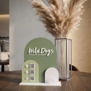 以下が含まれることがあります： 「Wild Dogs Professional Dog Training」の装飾看板。セージグリーンのアーチ型で、白い文字とソーシャルメディアのアイコンが付いています。看板は白いベースの上にあり、背景には乾燥したパンパスグラスの花瓶があります。