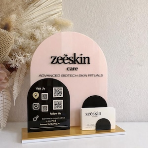Könnte beinhalten: Ein Displayständer mit einem rosa bogenförmigen Hintergrund und einer goldenen Basis. Der Ständer zeigt den Text "zeeskin care" und "ADVANCED BIOTECH SKIN RITUALS". Eine schwarze Tafel zeigt QR-Codes und Social-Media-Symbole.