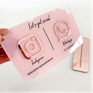 May include: A pink acrylic business card with rose gold Instagram and WhatsApp icons. The card has the text "Let's get social", "Instagram", "beautyloungewoerdennl", and "Whatsapp +31 6 87566955". A matching stand is also present.