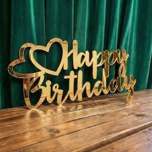Può includere: Un cake topper dorato "Happy Birthday" con un design a forma di cuore. La scritta è in corsivo. Il topper è posto su una superficie di legno davanti a una tenda di velluto verde.