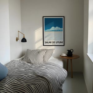 Op de afbeelding: Een wit bed met zwart-wit gestreept beddengoed en twee kussens. Een zwarte en gouden wandlamp is aan de muur achter het bed. Een kleine houten tafel met een zwarte kan en een witte mok staat naast het bed. Een ingelijste poster met de tekst "SALAR DE UYUNI Bolivia" hangt aan de muur boven de tafel.
