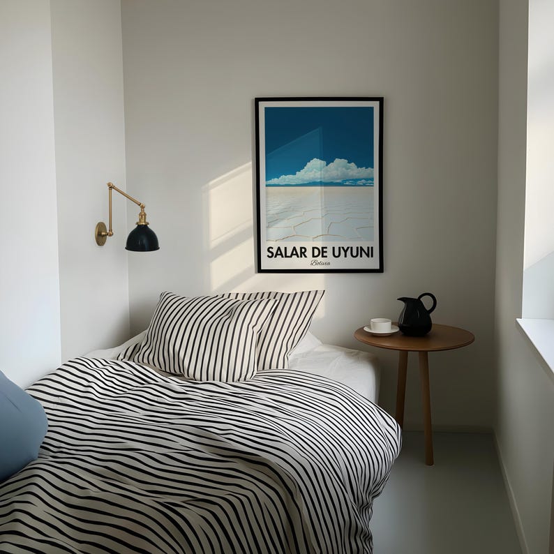Op de afbeelding: Een wit bed met zwart-wit gestreept beddengoed en twee kussens. Een zwarte en gouden wandlamp is aan de muur achter het bed. Een kleine houten tafel met een zwarte kan en een witte mok staat naast het bed. Een ingelijste poster met de tekst "SALAR DE UYUNI Bolivia" hangt aan de muur boven de tafel.