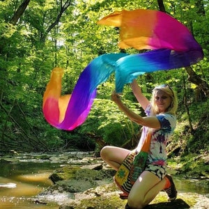 Puede incluir: Una mujer con una camisa de teñido anudado sostiene una bufanda de seda de colores arcoíris en un entorno boscoso. La bufanda ondea con el viento, creando una exhibición colorida. La mujer está arrodillada en una roca cerca de un arroyo.