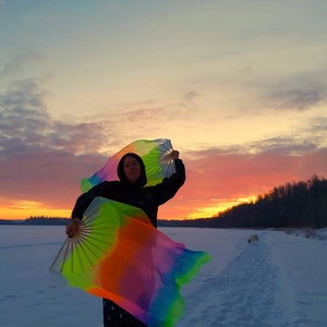 Peut inclure: Une personne portant une veste noire et tenant deux &eacute;ventails color&eacute;s, un dans chaque main, dans un champ enneig&eacute;. Les &eacute;ventails sont en tissu et ont un motif arc-en-ciel. La personne se tient devant un ciel de coucher de soleil.