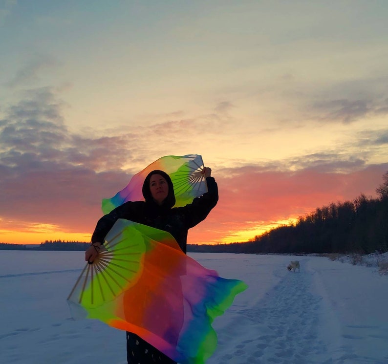 Peut inclure: Une personne portant une veste noire et tenant deux &eacute;ventails color&eacute;s, un dans chaque main, dans un champ enneig&eacute;. Les &eacute;ventails sont en tissu et ont un motif arc-en-ciel. La personne se tient devant un ciel de coucher de soleil.