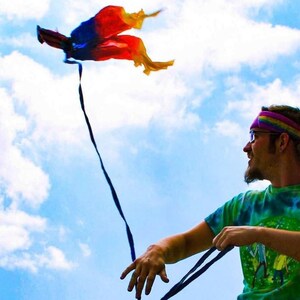 May include: A multi-coloured kite with red, blue, and yellow fabric panels and a long black tail flying against a bright blue sky with white clouds. The kite features a lightweight, flowing design that catches the wind, creating a vibrant display.