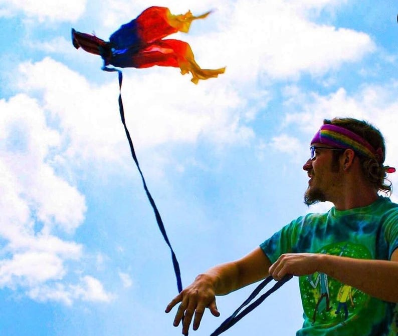 May include: A multi-coloured kite with red, blue, and yellow fabric panels and a long black tail flying against a bright blue sky with white clouds. The kite features a lightweight, flowing design that catches the wind, creating a vibrant display.